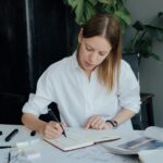 A woman in a white shirt reviewing technical drawings and notes at a desk with engineering tools, surrounded by plants and design materials.