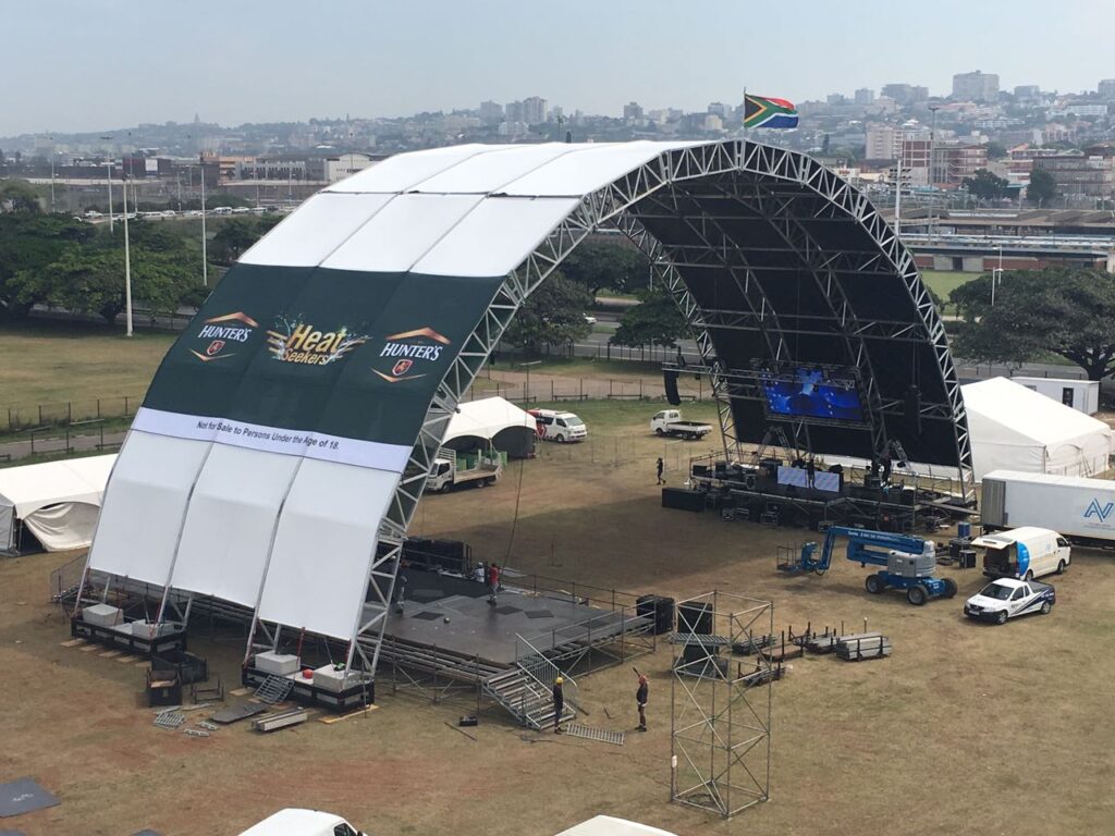 Aluminium trussing arch supporting a large stage with a curved roof, set up outdoors in an open field for an event, with cityscape background and Sout.