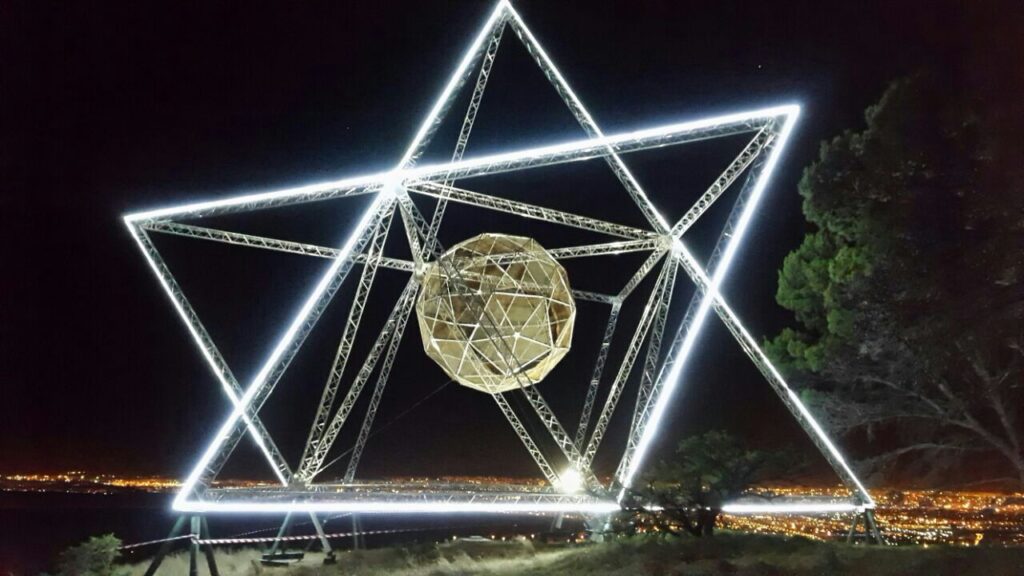 A large illuminated aluminium truss star structure with a central geometric sphere, used for event lighting and stage design at night.