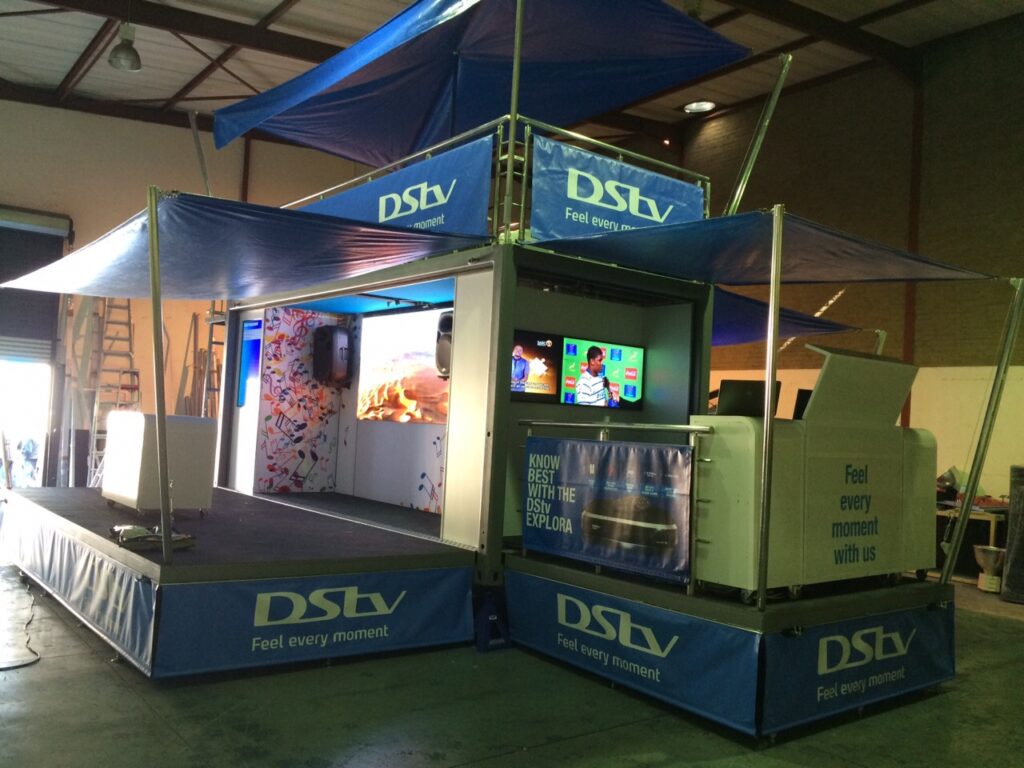Aluminium trussing structure with blue fabric canopies, supporting a promotional booth with screens and branding for event production and engineering.