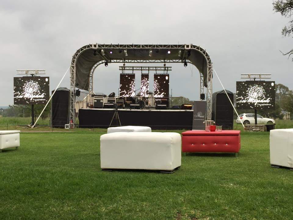 Aluminium trussing stage setup with lighting and sound equipment, outdoor event with seating, and a large LED screen backdrop on a grassy field.