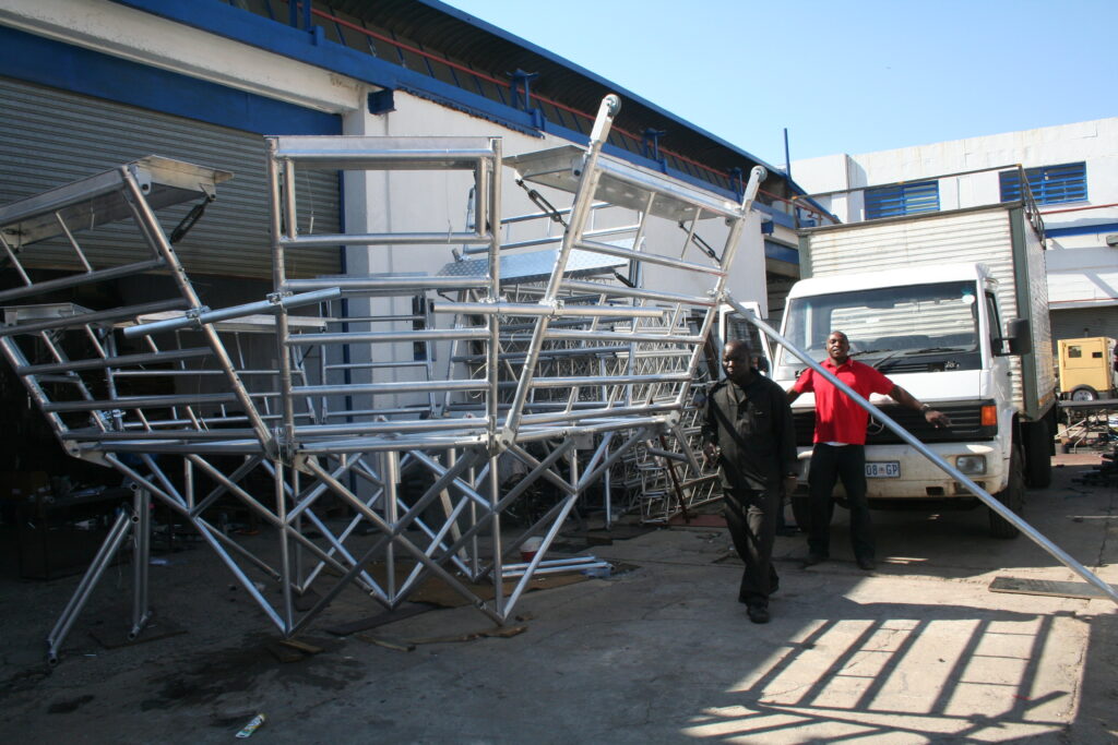 Aluminium trussing structure being assembled outdoors, with two workers handling components near a white van, suitable for event staging and rigging a.