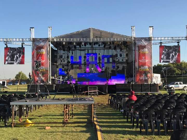 Aluminium trussing structure supporting stage lighting and audio equipment for outdoor concert setup, with seating area and event banners.