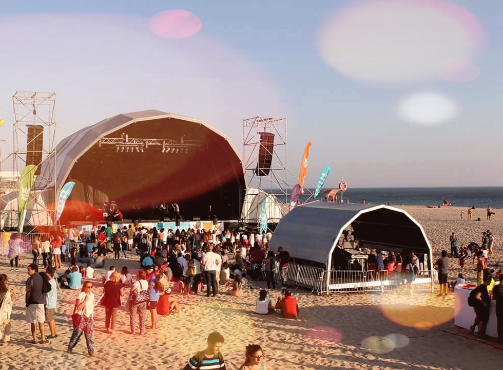 Aluminium trussing structure supporting a large outdoor stage at a beach event, with crowd and sound equipment, under a clear sky during sunset.