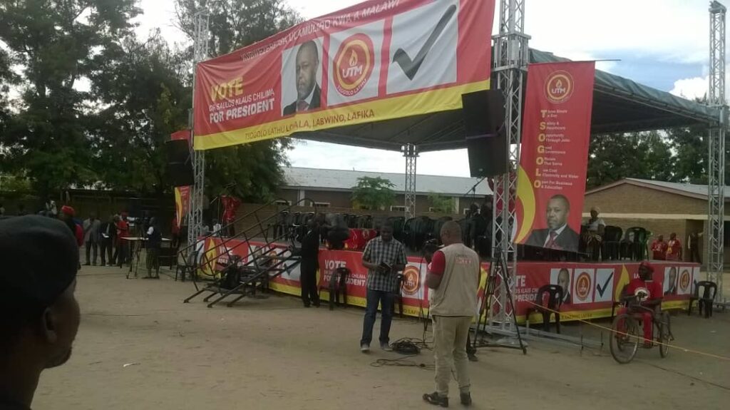 Aluminium trussing structure supporting a large political campaign banner at an outdoor event, with stage and audience in the background.