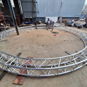 Aluminium trussing ring structure under construction in an industrial workshop, with workers assembling the framework for event or stage rigging.